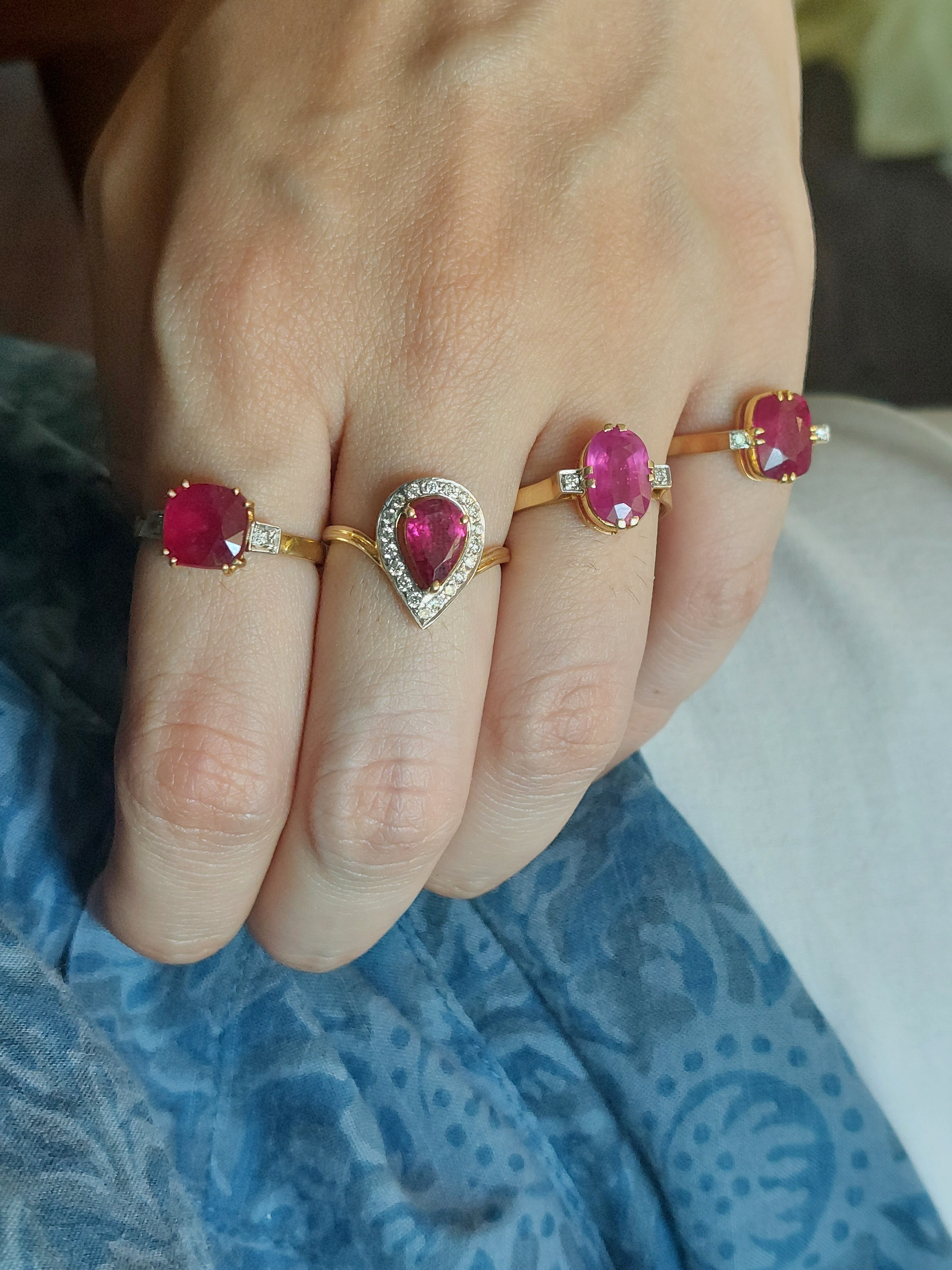 Four gold rings featuring vibrant pink gemstones in oval and pear shapes on a hand with a blue patterned fabric background.