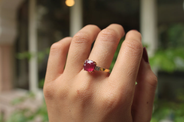 Gold ring with a vibrant oval red gemstone on a hand against a blurred outdoor background.